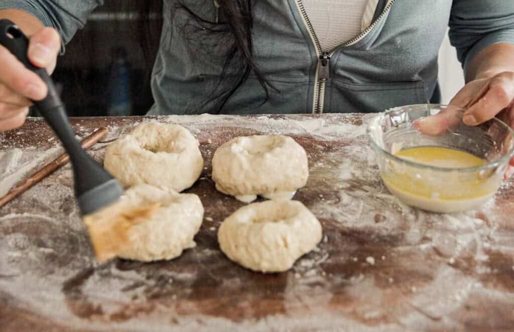 Person brushing wash onto four raw bagels on a floured wooden surface. A glass bowl with egg wash is nearby. The person is wearing a zip-up hoodie.